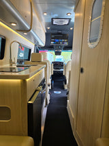Interior of an RV rental: a modern kitchen with a sink, cream seats, TV, and cockpit ahead, bathed in warm lighting.