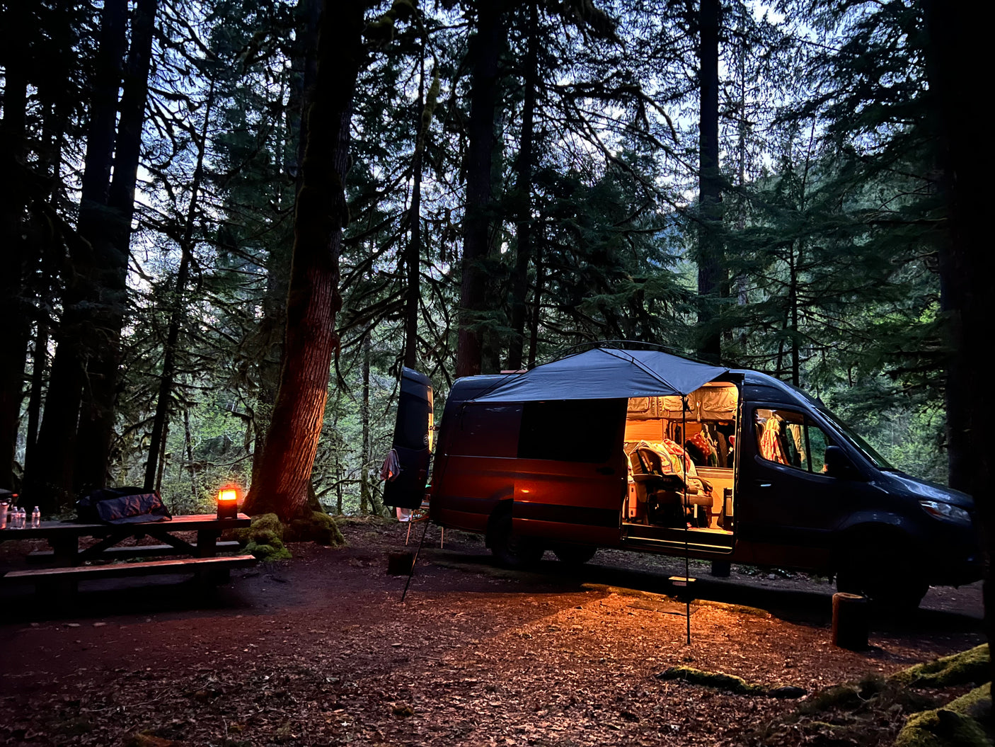 Cozy RV rental camper van parked in a dark forest campsite at dusk, with an illuminated interior and picnic table.