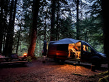 Cozy RV rental camper van parked in a dark forest campsite at dusk, with an illuminated interior and picnic table.