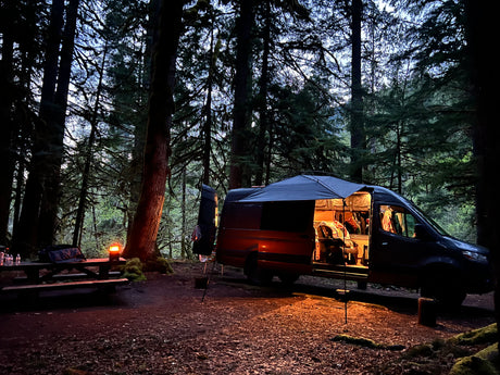 Cozy RV rental camper van parked in a dark forest campsite at dusk, with an illuminated interior and picnic table.