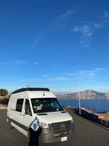 White RV rental van with roof rack at a scenic lake overlook. Mountains and blue sky ideal for road trips.