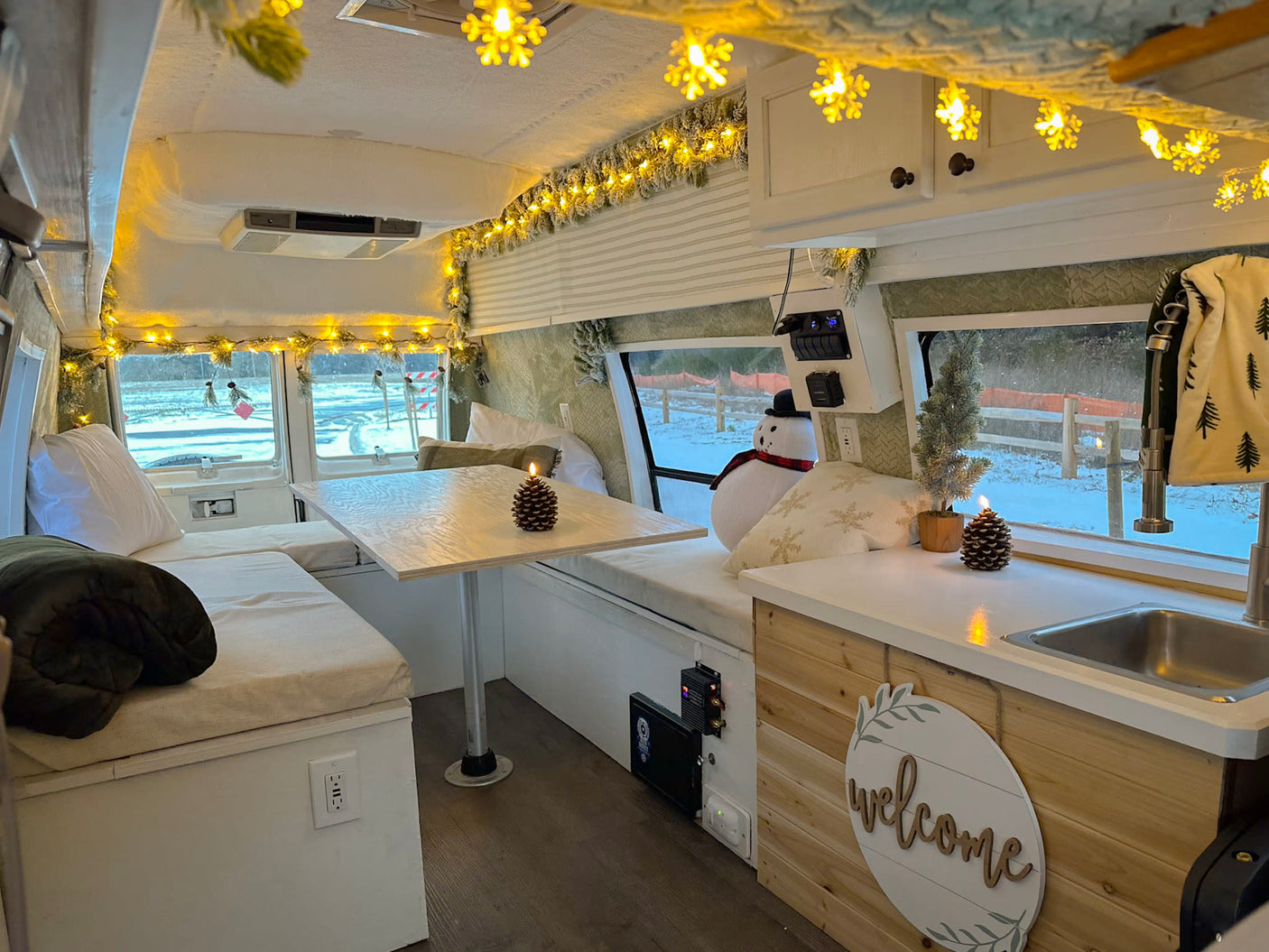 Cozy RV rental interior decorated with festive winter lights and accents. Features a dining area, kitchenette, and snowy outdoor views.