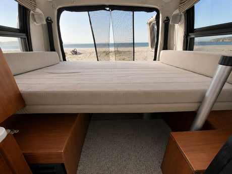 Inside an rv rental: A comfy bed provides a clear view of a sandy beach and ocean through the rear doors with mesh.