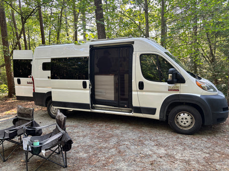 RV rental van, converted for camping, parked at a wooded site with chairs. Ready for adventure!