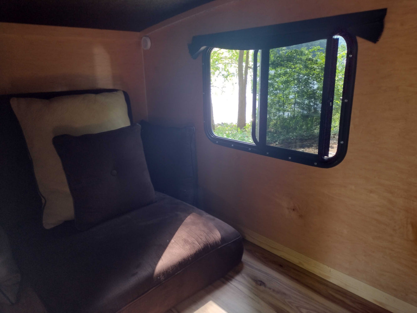 Cozy RV rental interior featuring a brown couch with pillows and a large window offering a view of lush trees and bright outdoors.
