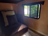 Cozy RV rental interior featuring a brown couch with pillows and a large window offering a view of lush trees and bright outdoors.