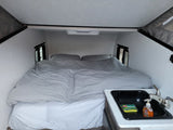 Inside an rv rental: a light-colored interior with a large bed, grey bedding, small windows, and a compact sink with soap.