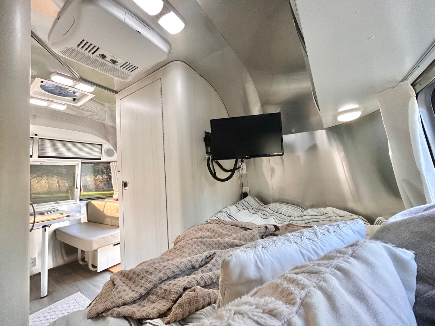 A bright, modern rv rental interior. The bedroom features a cozy bed with throws & a TV. A small dinette/kitchen area is visible through the doorway. — photo 9