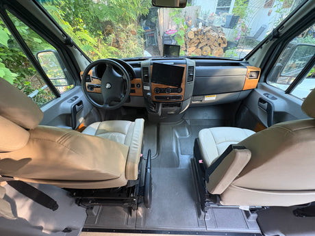 Interior view of an RV rental showing front seats, dashboard with screens, and an outdoor scene through the windshield.