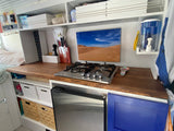 Inside an RV rental, a compact kitchen with wood-look counter, built-in stovetop, fridge, and storage. Perfect for road trips.