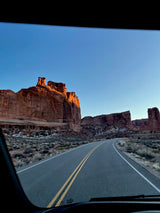 RV rental driving through a red rock desert. Towering, sunlit formations line a winding road. A clear view from inside the vehicle.