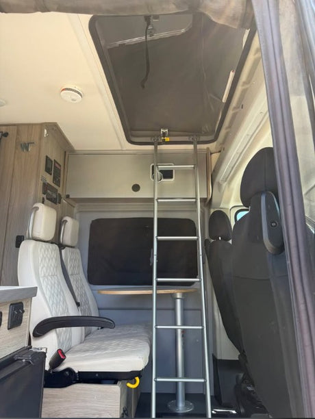 RV rental camper van interior. Ladder to upper bed, with white quilted seats and a small table below.