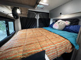 Comfortable RV rental bedroom: cozy bed with colorful bedding, fan, overhead storage, and privacy window covers.