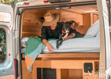 Inside a comfy RV rental: A person and dog relax on the bed of a wooden camper van. Ideal for travel life.