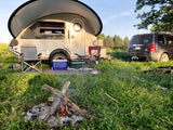Perfect RV rental camping setup with a teardrop trailer, chairs, cooler, and a warm campfire at sunset.