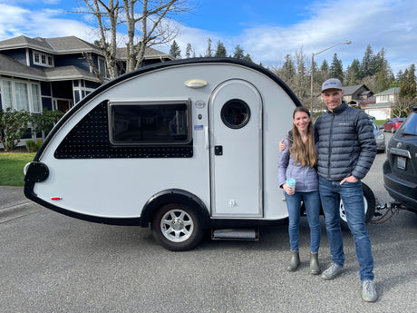 A smiling couple poses with their gray T@B rv rental teardrop trailer, ready for travel & adventure.