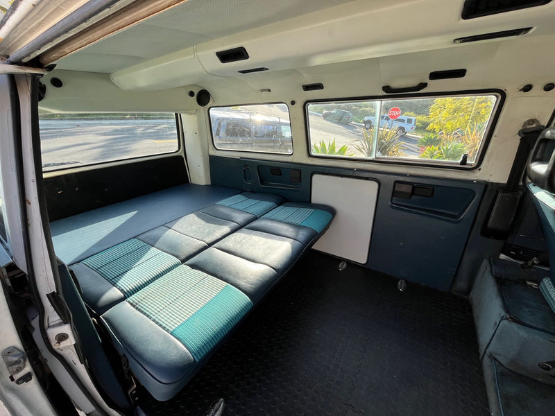 Comfortable RV rental interior: Blue/teal checkered bed, dark floor, and windows showing the street. — photo 9