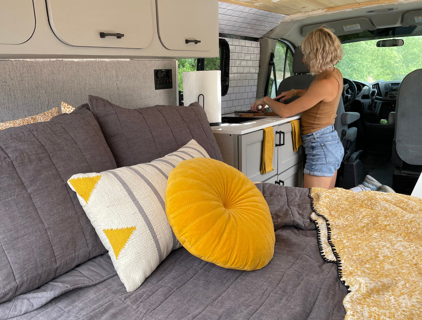 This RV rental shows a woman cooking in a compact kitchen, with a cozy bed featuring gray and yellow pillows in the foreground. — photo 14