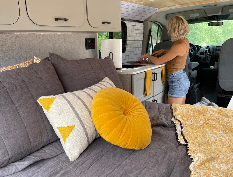This RV rental shows a woman cooking in a compact kitchen, with a cozy bed featuring gray and yellow pillows in the foreground. — photo 14