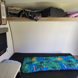 Cozy rv rental sleeping area with a colorful sleeping bag, overhead luggage, linen storage, and a small table.