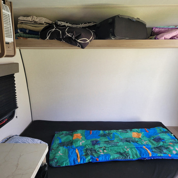 Cozy rv rental sleeping area with a colorful sleeping bag, overhead luggage, linen storage, and a small table.