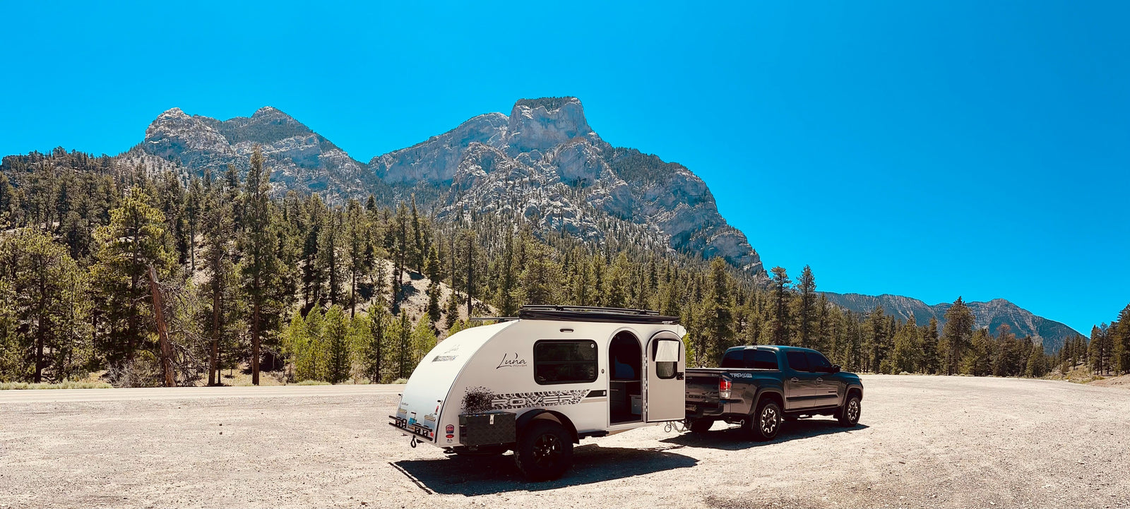 A Tacoma truck tows a compact teardrop RV rental, parked in a scenic mountain and forest landscape. Ready for adventure!