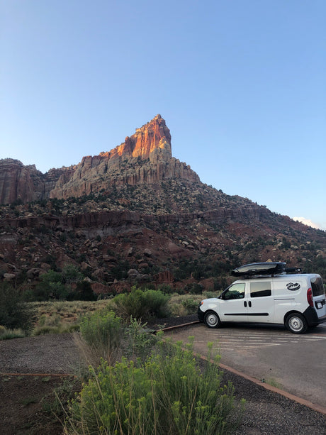 White camper van (RV rental) with roof box parked before a majestic, sunlit mountain. Ideal for desert road trips & camping.