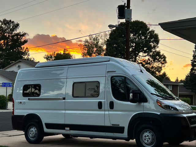 White Winnebago Solis, a camper van suitable for an RV rental, parked on a street at sunset with vibrant orange clouds.