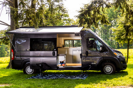 Stylish dark gray RV rental camper van, side door open to reveal a modern kitchen, with awning, chairs, & mat on grass.
