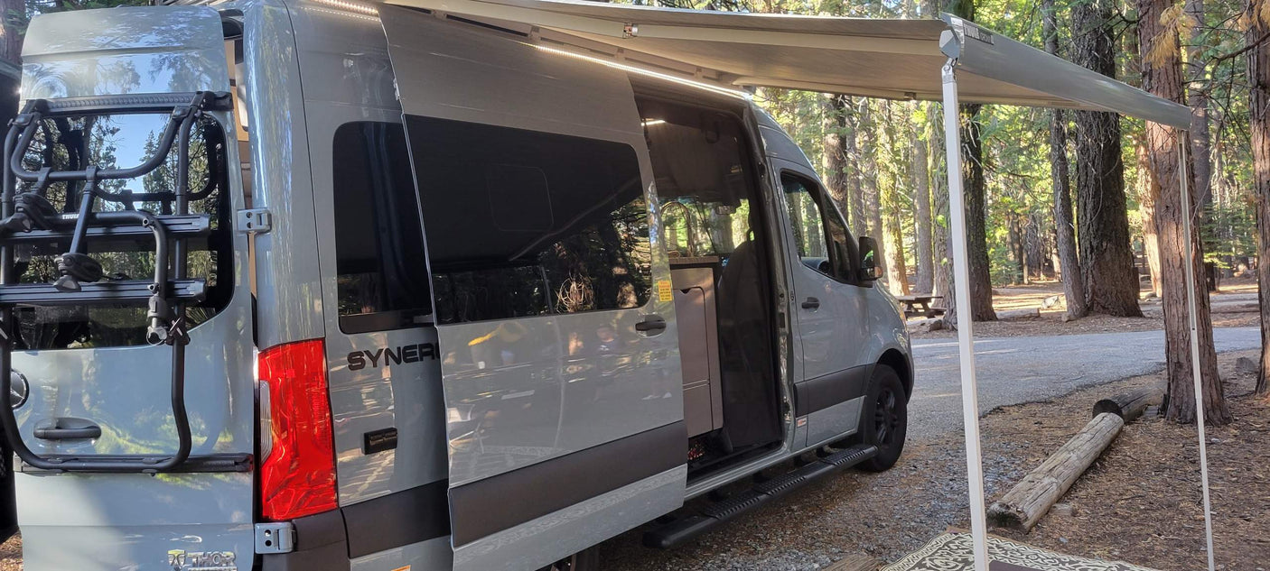 A grey Thor Synergy camper van with extended awning & bike rack at a forest campsite. Ideal for an RV rental experience.
