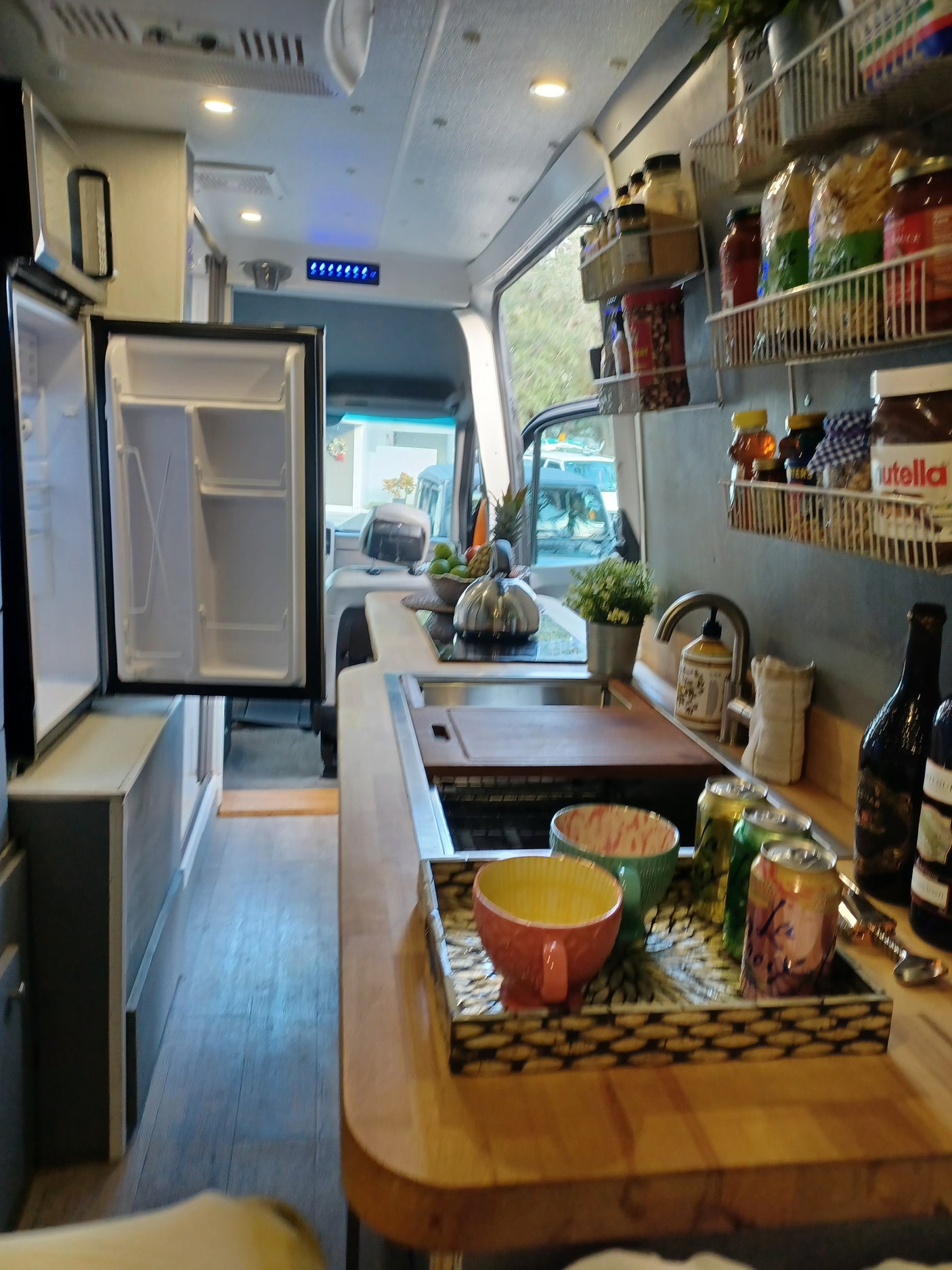 RV rental kitchen: open fridge, organized shelves with food, sink, and countertop with fruit & mugs. Ready for adventure!