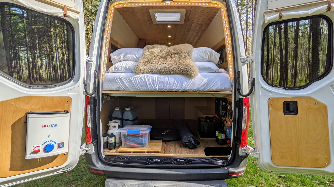 Open rear of an RV rental camper van, revealing a comfy bed, gear storage, HOTTAP water heater, and forest beyond.