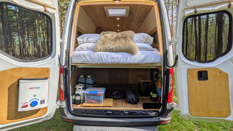 Open rear of an RV rental camper van, revealing a comfy bed, gear storage, HOTTAP water heater, and forest beyond.