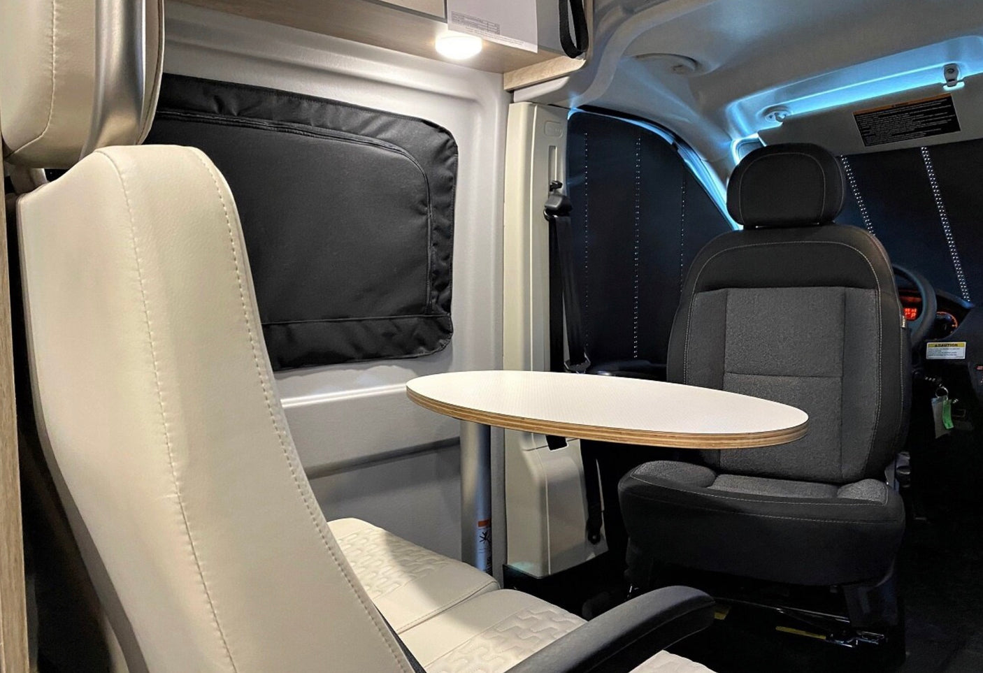 This **RV rental interior** features a cream swivel seat, an oval table, and the driver's seat, with black window shades.