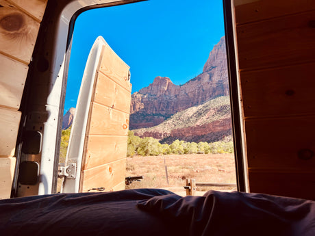 RV rental view: Majestic red mountains, green trees, & field through open doors of a cozy, wood-paneled van.