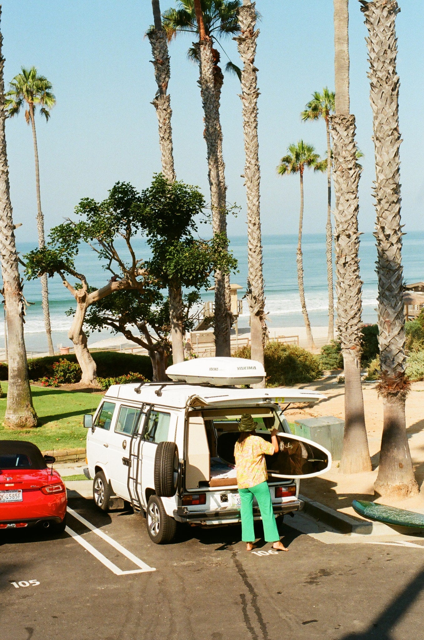 A white RV rental van with surfboards is parked on a sunny road near palm trees and the ocean. A person handles gear. — photo 8