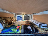 This RV rental offers a cozy sleeping area. Features include wood paneling, a patterned quilt, and bright windows.