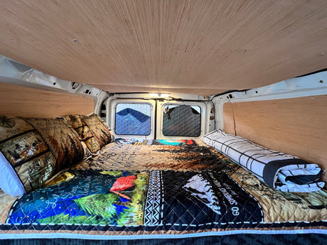 This RV rental offers a cozy sleeping area. Features include wood paneling, a patterned quilt, and bright windows.
