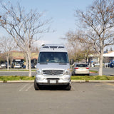 A silver Mercedes Sprinter van, resembling an RV rental, parked head-on in a bright outdoor lot with bare trees.