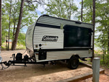 This Coleman travel trailer RV rental is parked on a dirt campsite among trees, with a lake visible in the background.