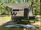 RV rental: A white and black travel trailer with an extended awning is set up in a shaded forest campsite.