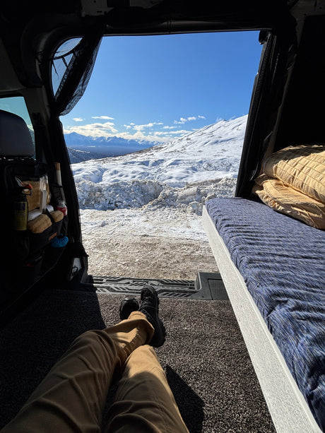 View from an RV rental's rear, showing a person's legs, interior bed, and a breathtaking snowy mountain landscape.