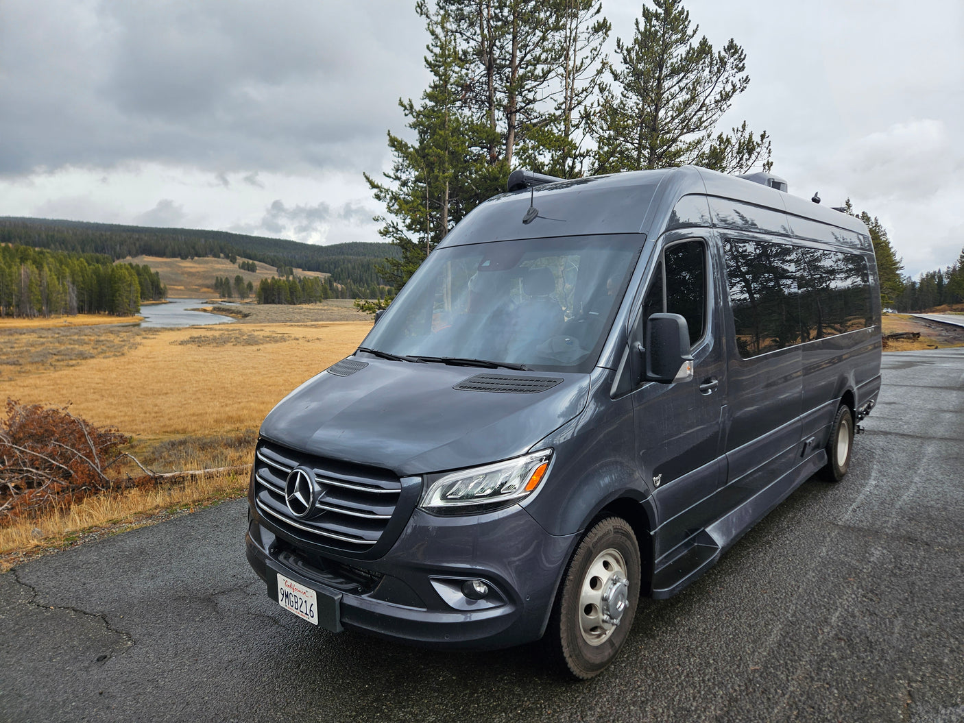 A sleek grey Mercedes-Benz Sprinter rv rental parked by a winding river, golden plains, and evergreen forests under a cloudy sky.