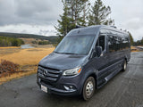 A sleek grey Mercedes-Benz Sprinter rv rental parked by a winding river, golden plains, and evergreen forests under a cloudy sky.