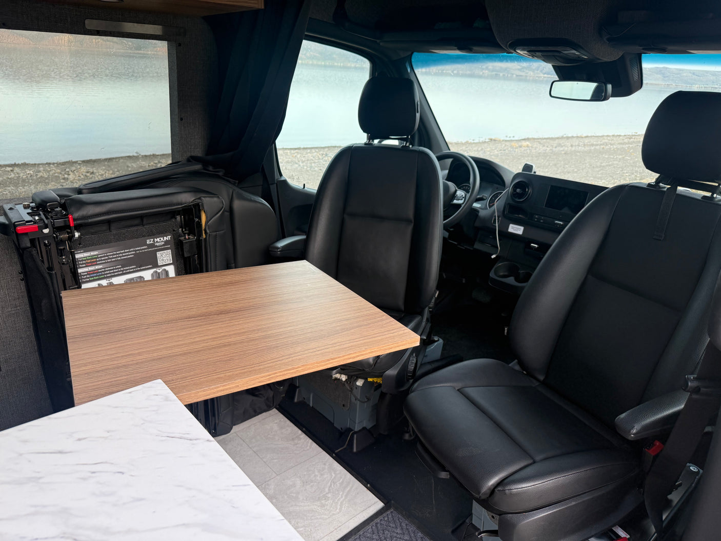 RV rental interior featuring swiveling driver and passenger seats with a deployed table, offering a scenic lake view.