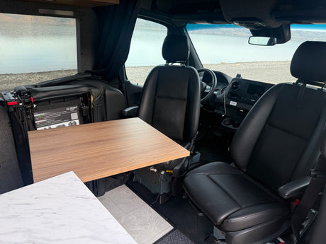 RV rental interior featuring swiveling driver and passenger seats with a deployed table, offering a scenic lake view.