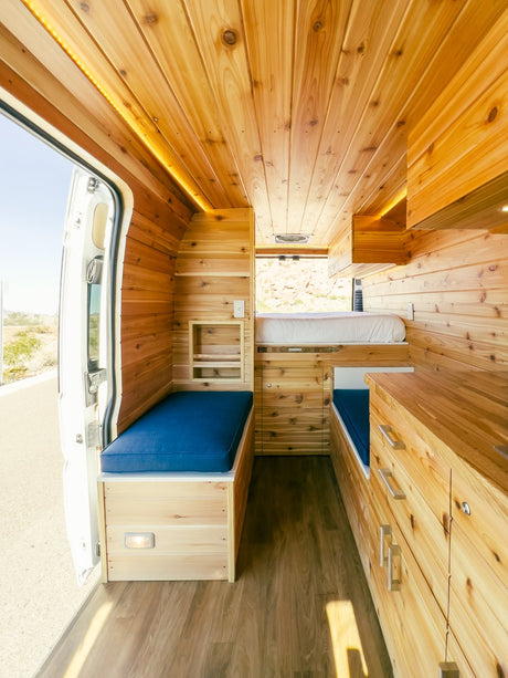 Inside a warm, custom wood camper van, an ideal RV rental. Features a high bed, blue bench, and built-in storage units.