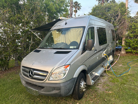 Silver Mercedes Sprinter RV rental parked on grass with hookups connected, awning extended.