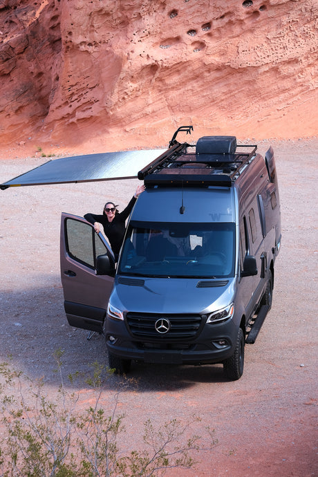 Adventure awaits! Smiling woman beside an RV rental van with an awning, parked against a red rock desert. Ideal for travel.