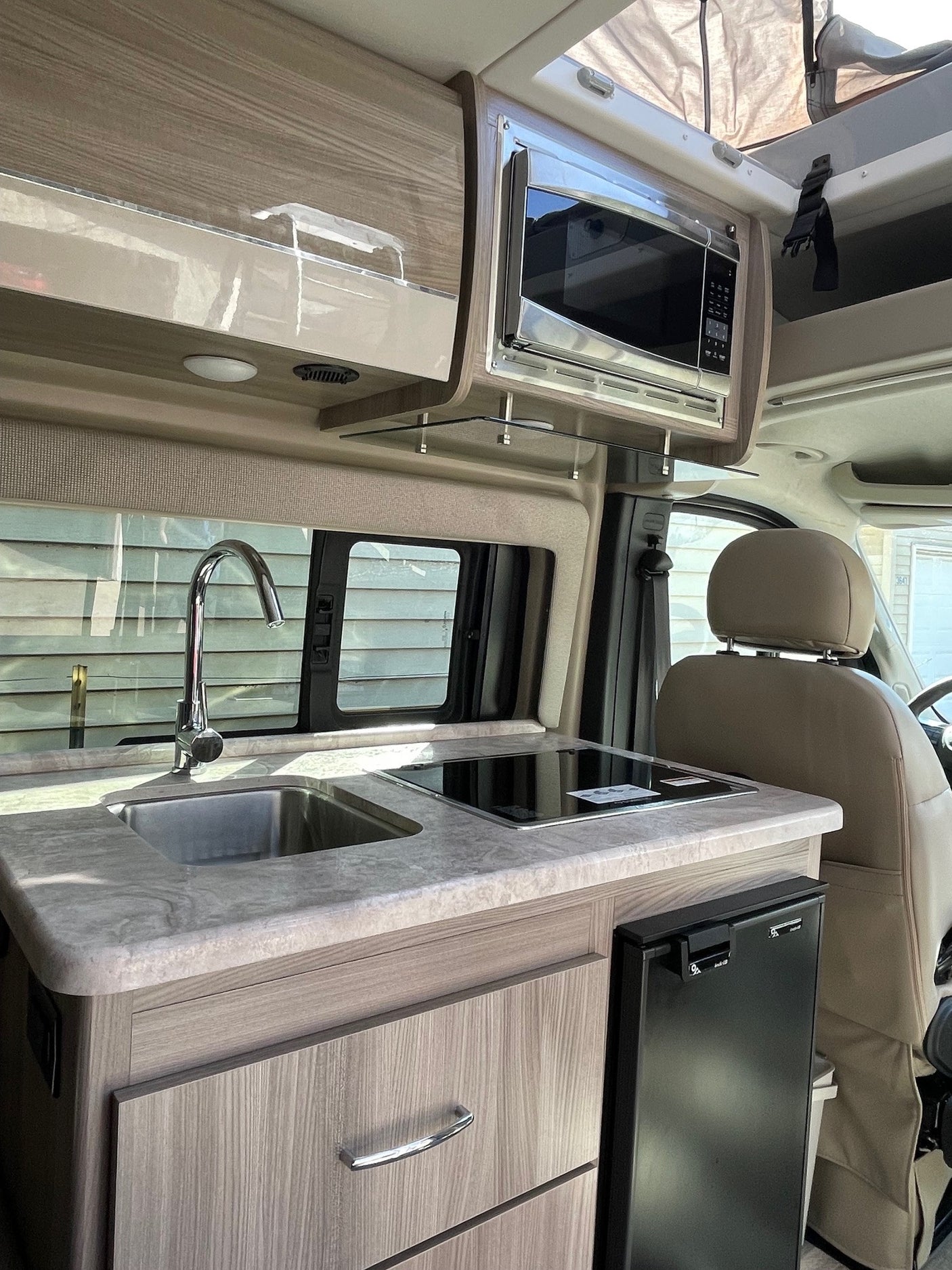 Inside an RV rental: a modern galley with a sink, microwave, induction cooktop, fridge, and wood-grain cabinets.